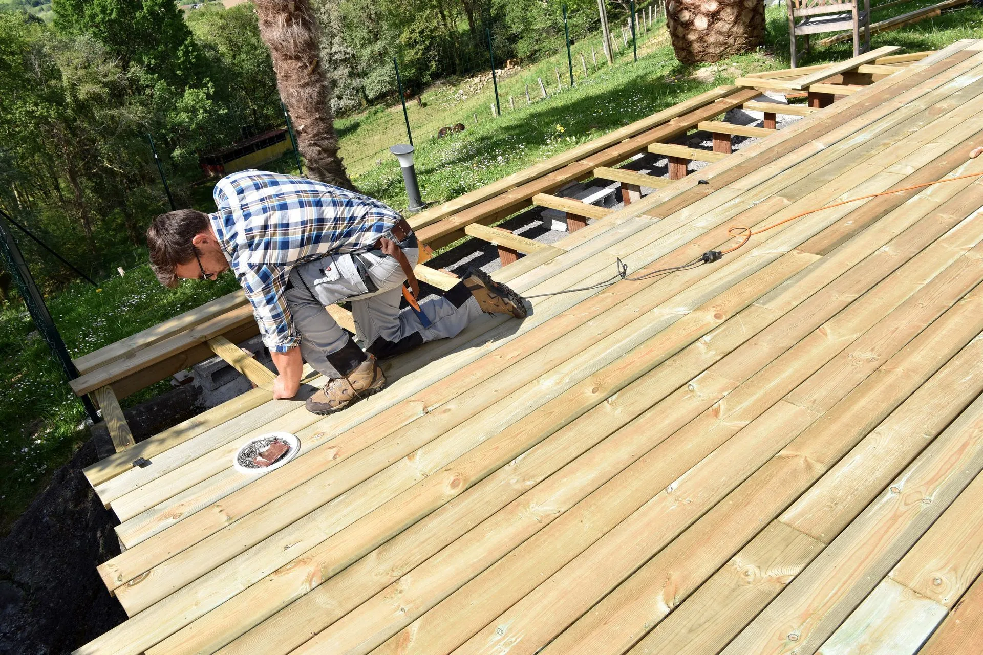 Worker installing wooden deck boards on an outdoor deck frame in a wooded area.