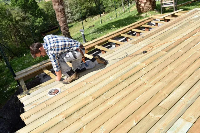 Worker installing wooden deck boards on an outdoor deck frame in a wooded area.