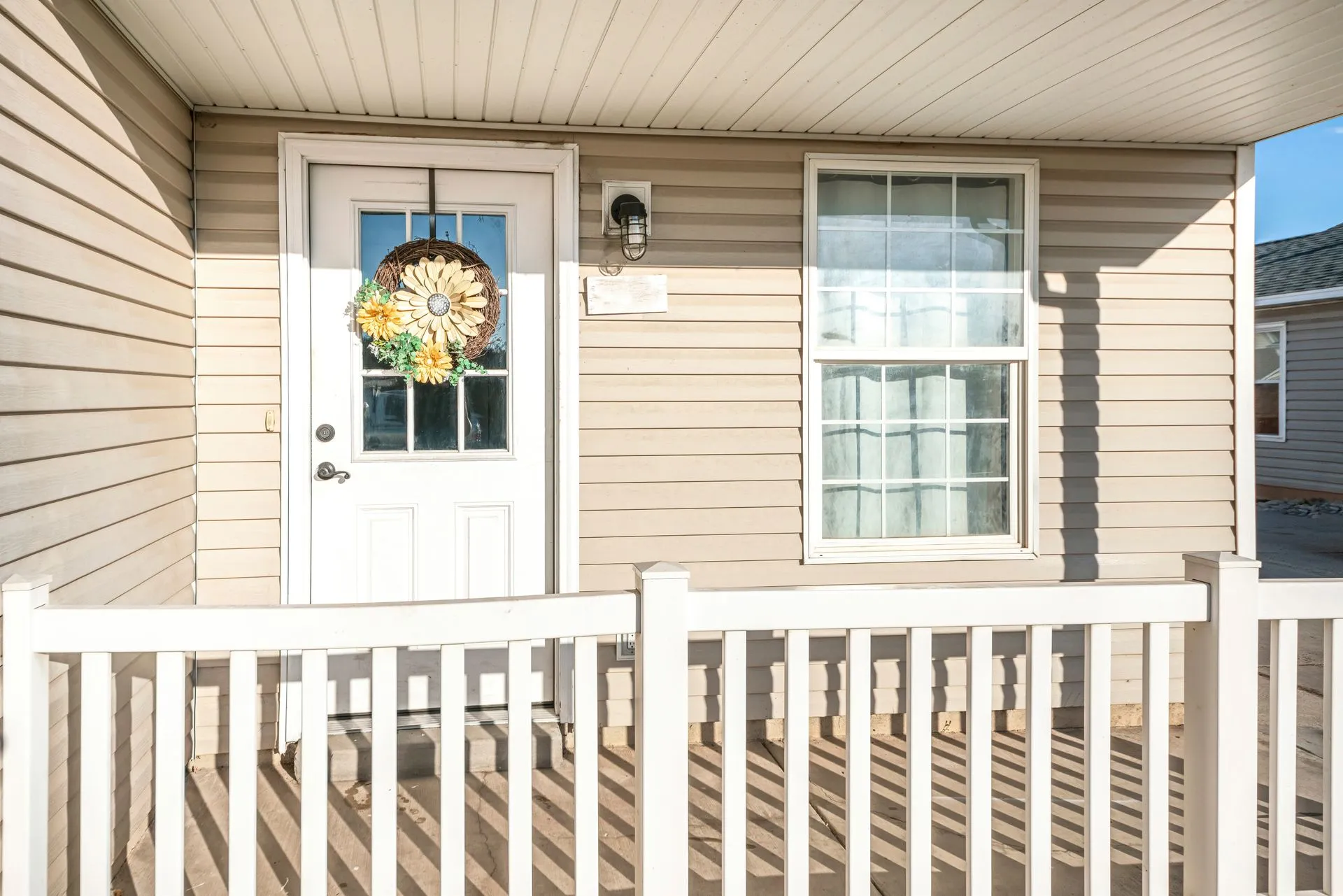 Front porch with white railing, beige siding, white door with wreath, and a window beside it